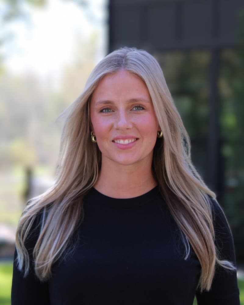 Kerigan Morris of TKO Homes smiling outdoors in a black top, with trees and a dark modern home behind her